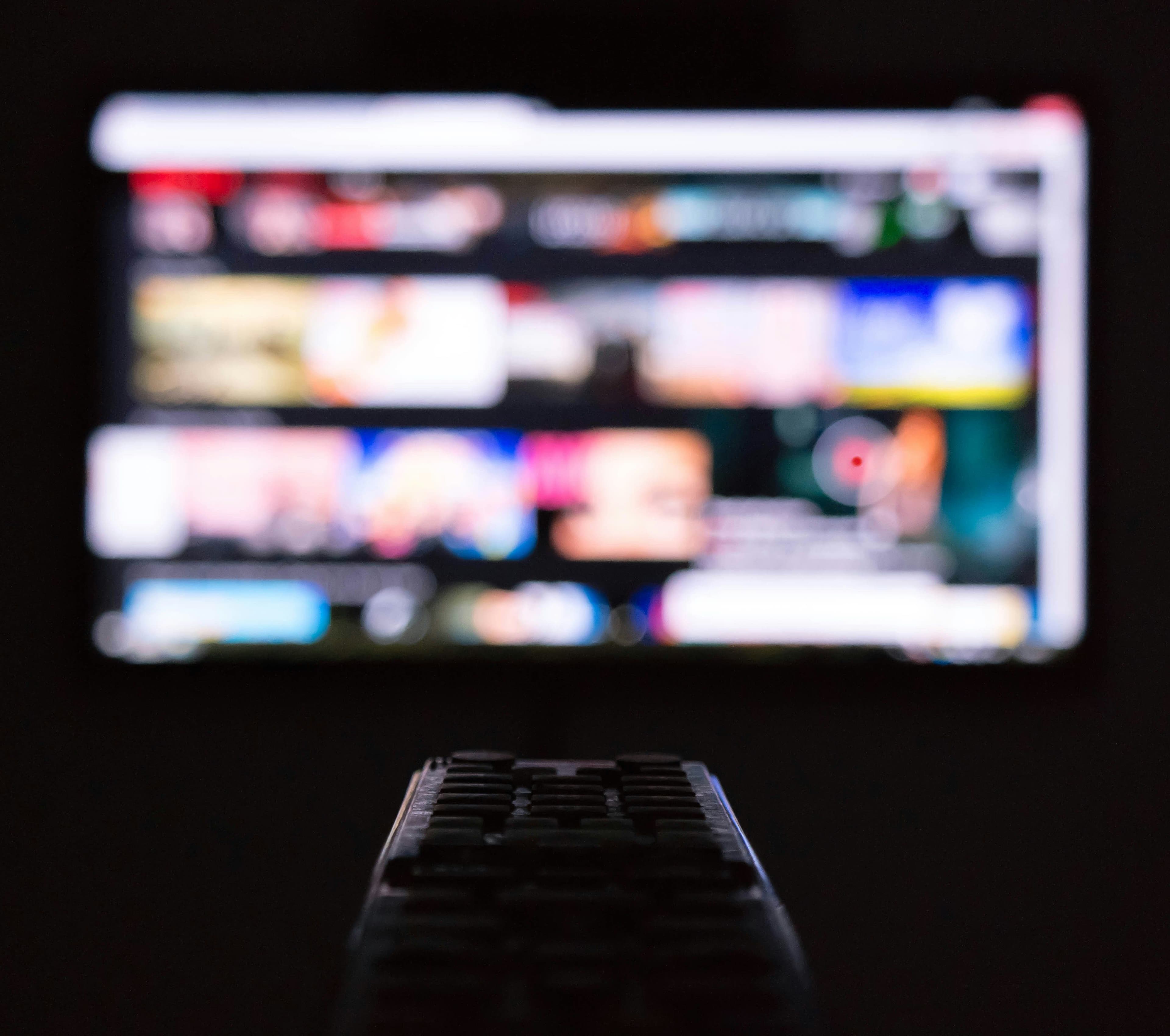 Fernbedienung vor einem unscharfen Smart-TV-Bildschirm mit mehreren Kacheln und Menüelementen.
